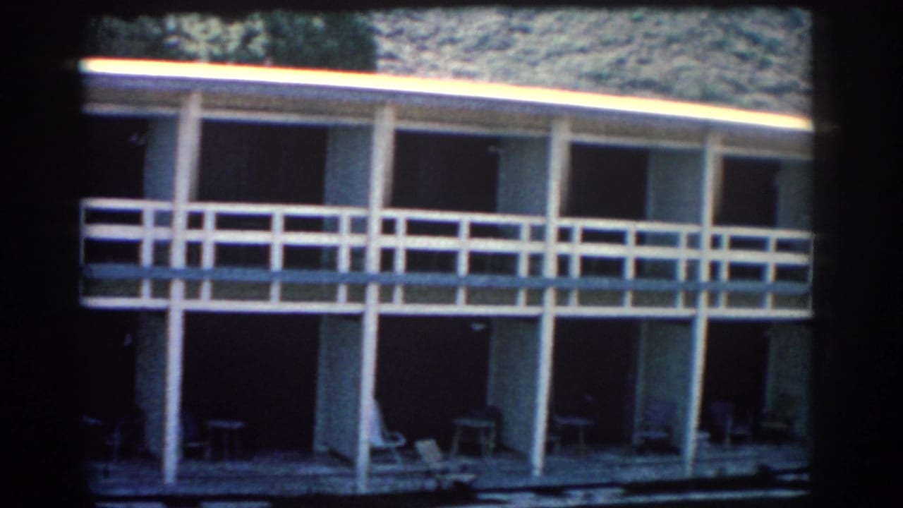 1962 Rhodes home-movie frame showing a modernist two-story building with white railings, shaded doorways, and patio chairs.