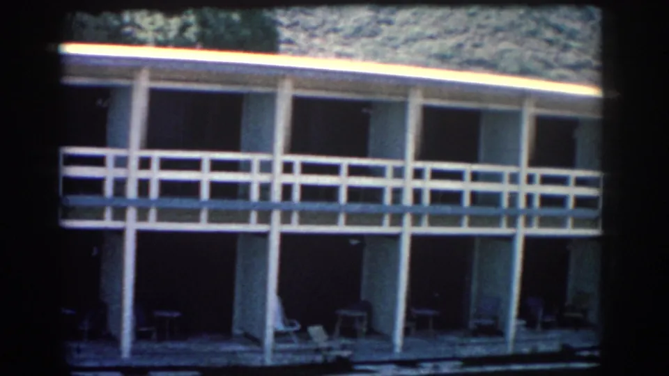 1962 Rhodes home-movie frame showing a modernist two-story building with white railings, shaded doorways, and patio chairs.