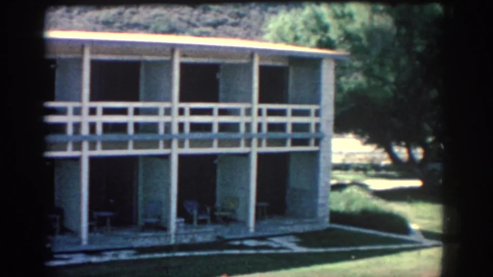 1962 Rhodes travel frame showing the building as the pan reveals more lawn, trees, and sunlit grounds beside it.