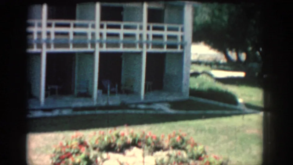 1962 Rhodes home-movie frame with the building, lawn, trees, and a flower bed visible during the rightward pan.