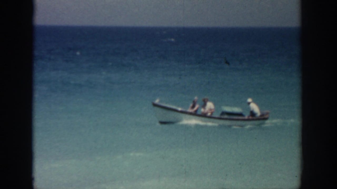 1962 Rhodes, Greece 8mm frame showing a small boat with passengers moving across turquoise water near shore.
