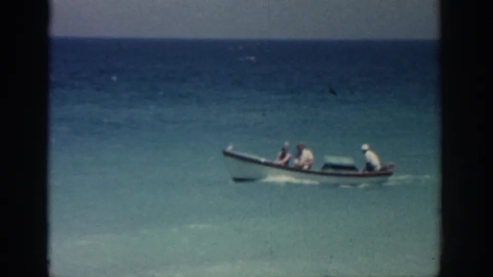 1962 Rhodes, Greece 8mm frame showing a small boat with passengers moving across turquoise water near shore.