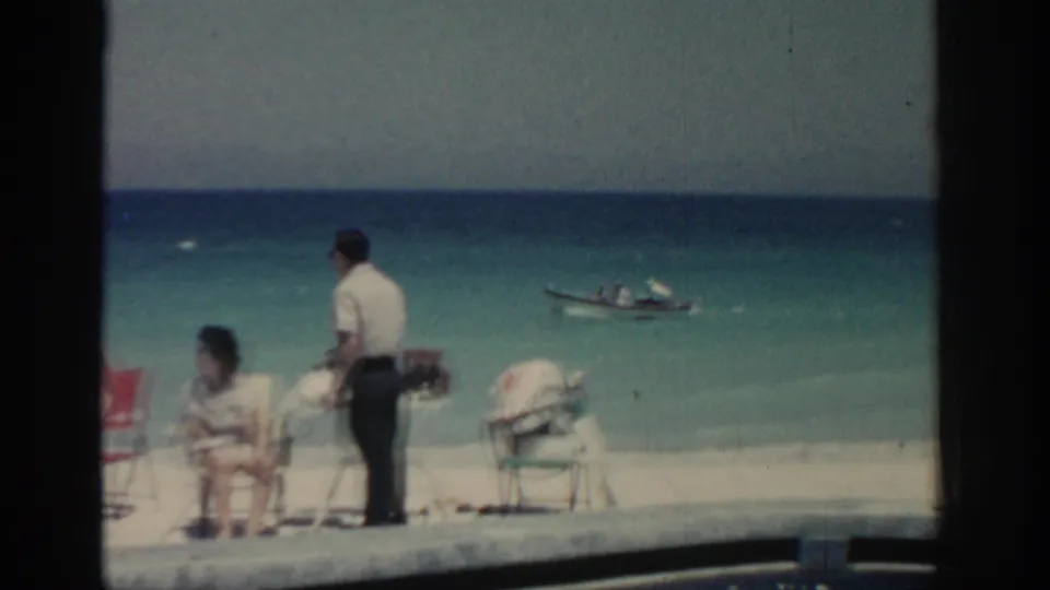 Adults relax on a Rhodes beach while a man stands near chairs and a small boat continues offshore.