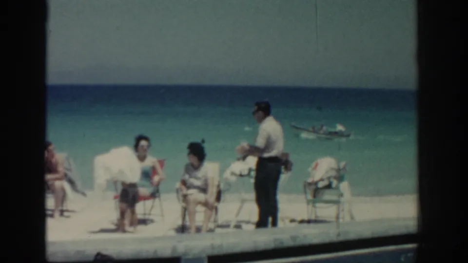 1962 Rhodes beach scene with seated adults, a standing man, folding chairs, and a small boat in the background.