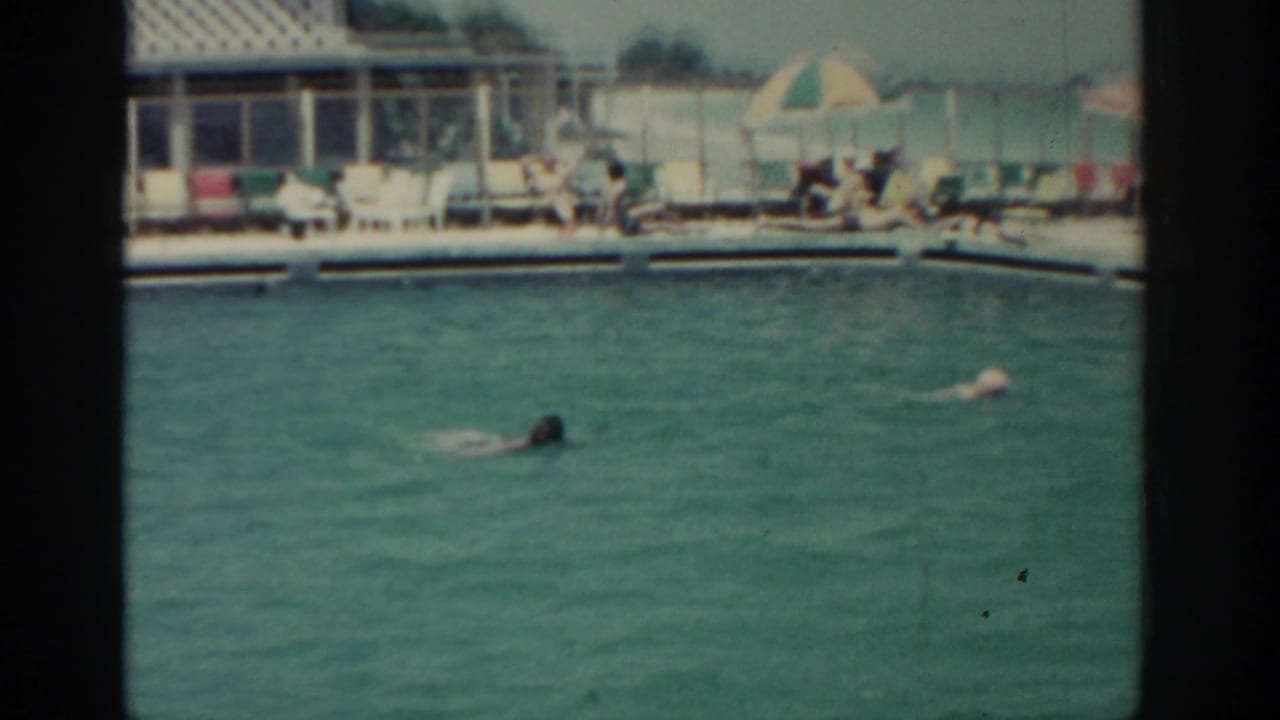 1962 8mm home-movie view of two people swimming in a resort pool in Rhodes, Greece, with guests seated under umbrellas beyond the water.
