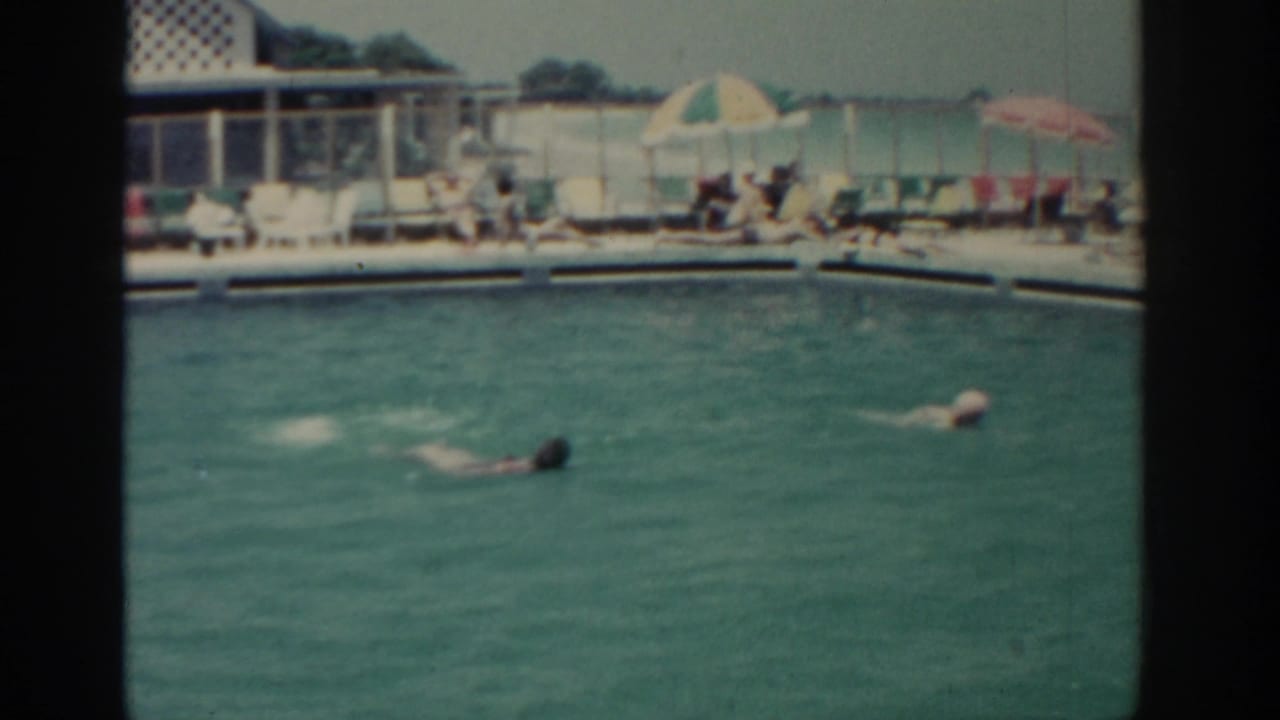 Two distant swimmers are seen in a Rhodes resort pool, with lounge chairs, poolside guests, and colorful umbrellas visible behind them.
