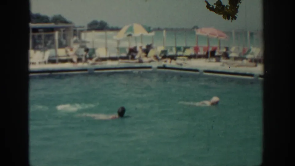 A soft 8mm frame shows two swimmers in a Rhodes resort pool, with lounge chairs, umbrellas, and seated guests along the far side.