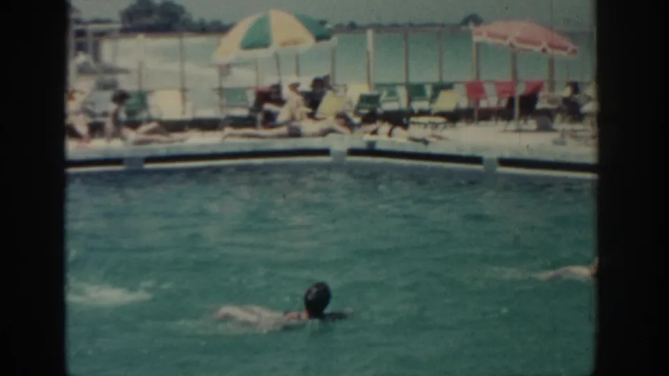 In this 1962 Rhodes poolside frame, one swimmer is closer to camera while another is partly visible at right, with guests and umbrellas behind them.