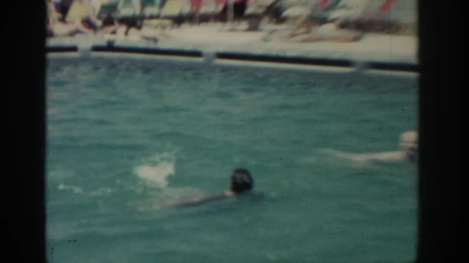 1962 Rhodes home-movie frame with swimmers in a resort pool and blurred poolside lounge chairs behind them.