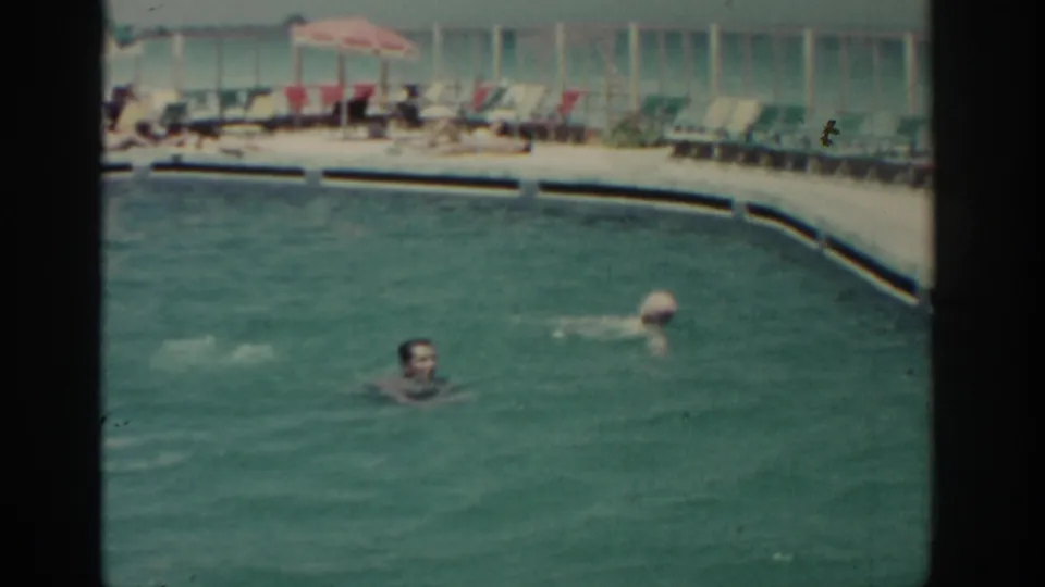 Wide 1962 Rhodes resort-pool view with swimmers in turquoise water and rows of lounge chairs beneath umbrellas.