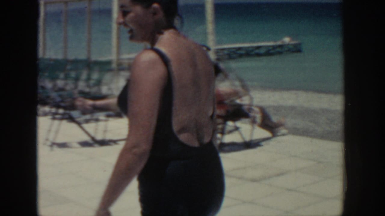 In Rhodes, Greece in 1962, a woman in a black one-piece swimsuit walks past lounge chairs on a bright pool deck.