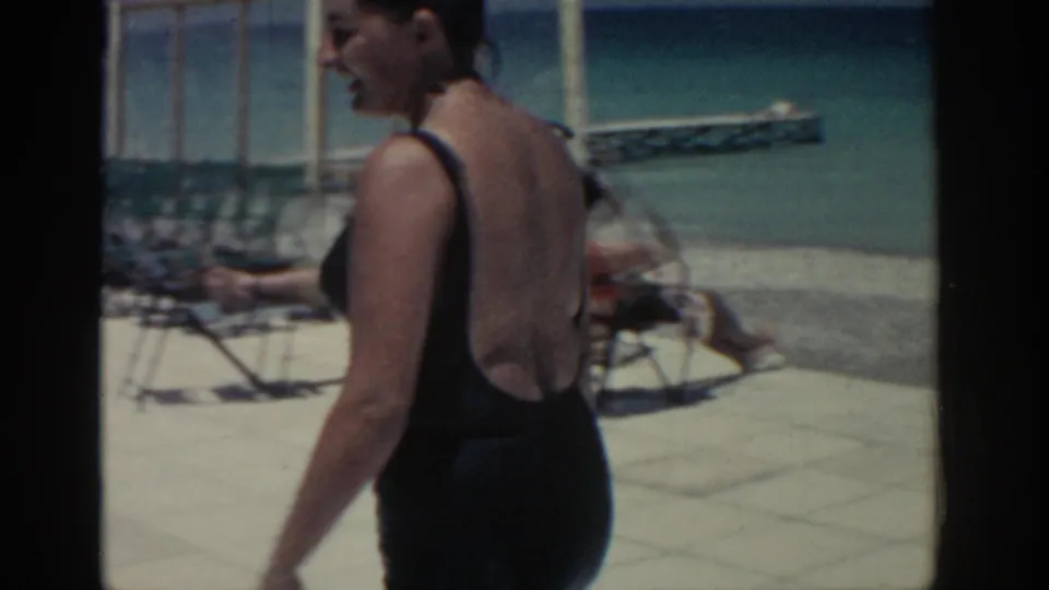 In Rhodes, Greece in 1962, a woman in a black one-piece swimsuit walks past lounge chairs on a bright pool deck.