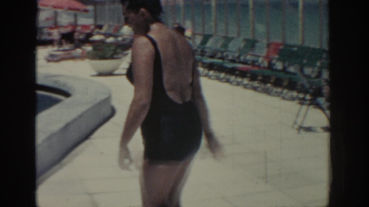 The woman continues along the Rhodes resort pool edge, with rows of lounge chairs and umbrellas visible behind her.