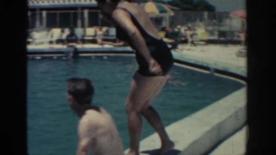 At the Rhodes pool in 1962, the woman begins her jump from the edge while another adult sits in the water nearby.