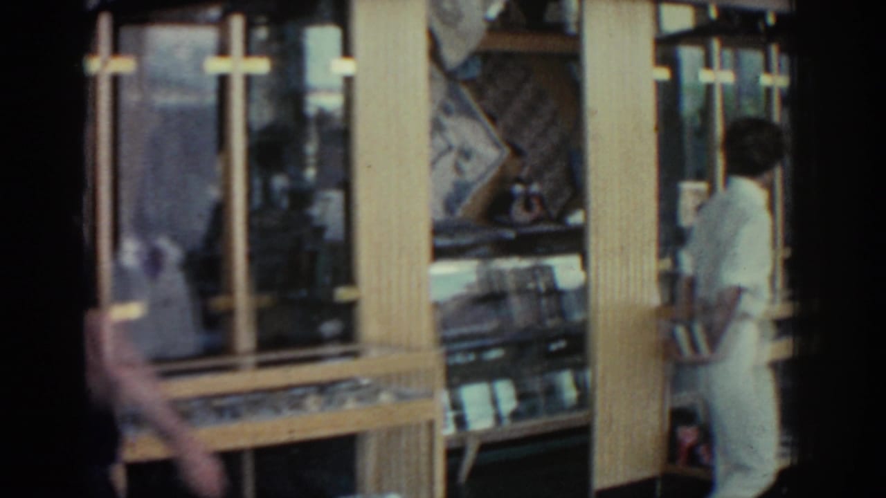 A dark-clothed figure crosses the foreground while gold-framed display cases remain visible in the Rhodes shop interior.