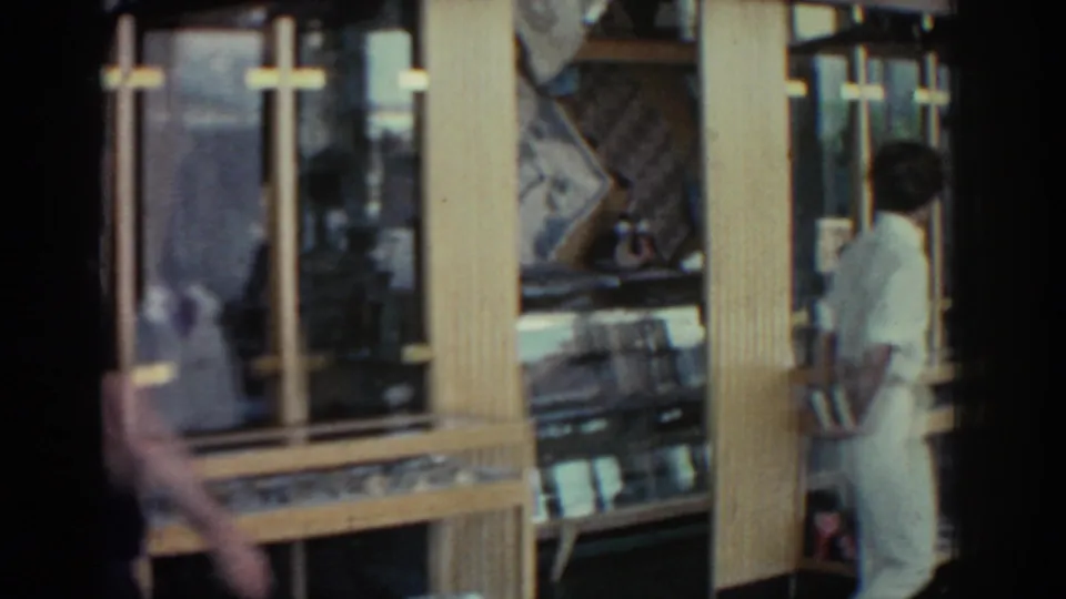 A dark-clothed figure crosses the foreground while gold-framed display cases remain visible in the Rhodes shop interior.