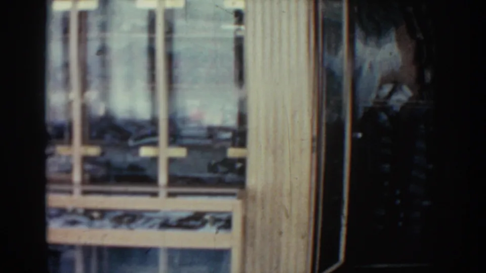 1962 Rhodes home-movie still showing the lower section of a wooden window, glass reflections, pale trim, and indistinct interior objects.