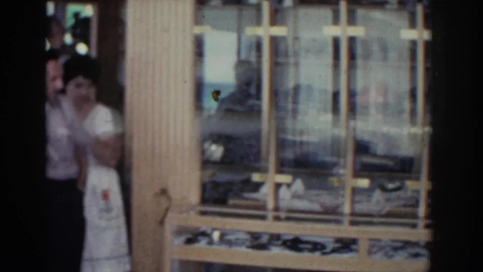 A man and woman appear in a shop doorway in Rhodes, framed by wooden windows and interior displays.