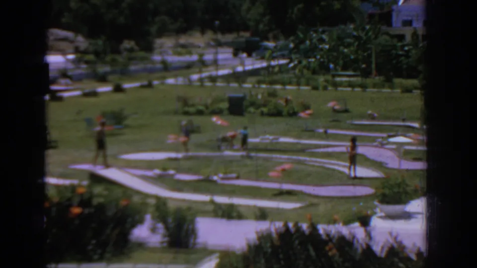 A landscaped miniature golf course in Rhodes appears with grass, winding paths, plants, and small figures playing or walking.