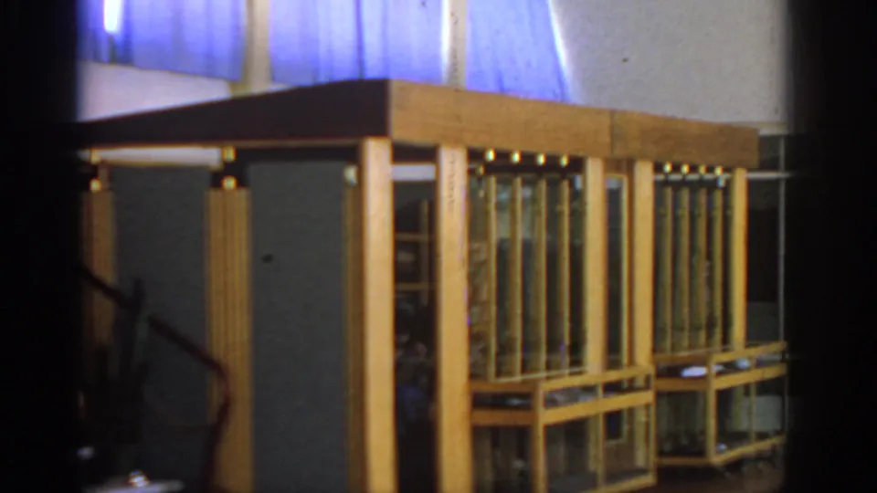 1962 hotel lobby detail in Rhodes, with wood-and-glass cases, blue curtains, and a shadowed stair rail at the left edge.