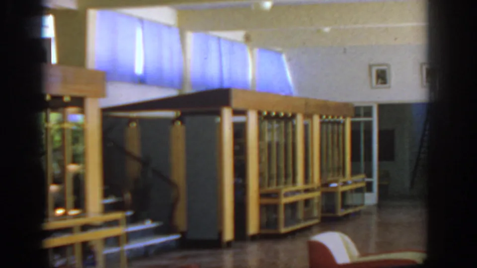 Wide 1962 Rhodes hotel interior showing display counters, blue-curtained windows, wall portraits, polished floor, and a red chair.