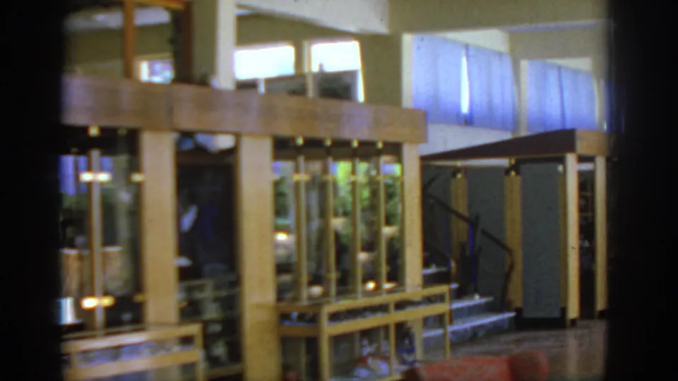 1962 Rhodes home-movie frame closer to the wooden display case, with glass panels, bright high windows, polished floor, and a stairway behind it.