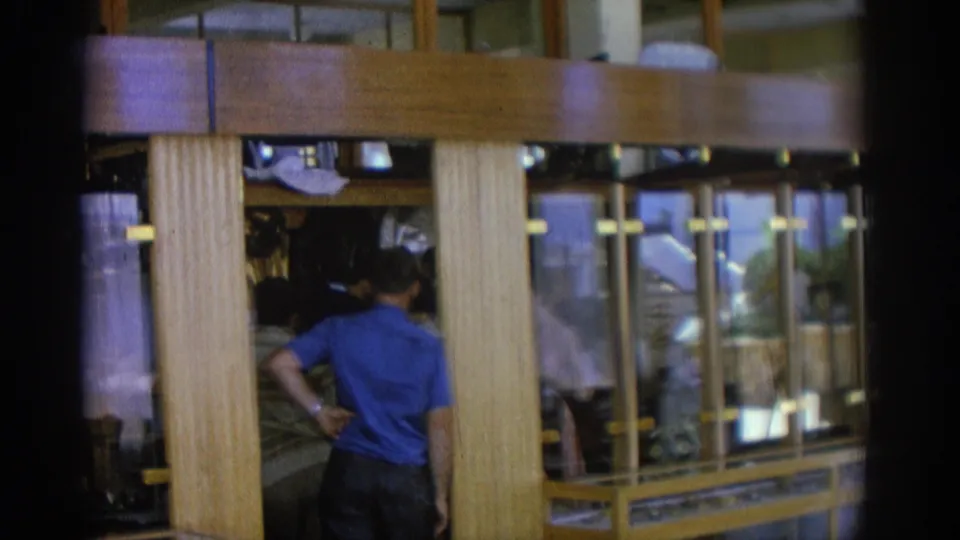 The camera continues past a man seen from behind beside glass-front cases in the 1962 Rhodes beachfront restaurant interior.