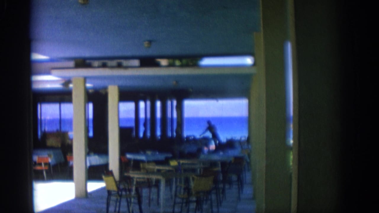 Restaurant interior with white pillars, tables, chairs and blue sea visible through large windows.