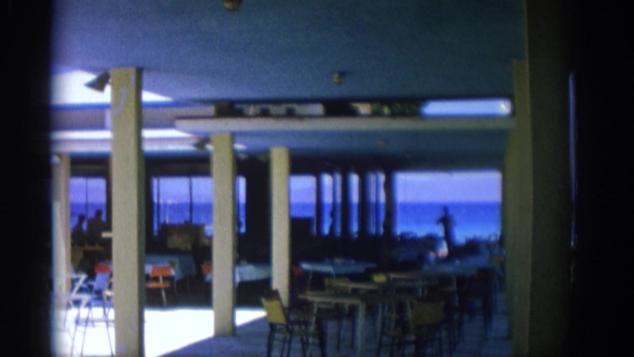 Sunlit restaurant seating area with columns, colorful chairs and ocean visible through the windows.