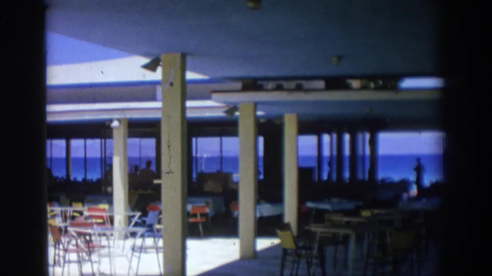 1962 Rhodes home-movie frame of a shaded oceanside dining room with white pillars, café chairs and the sea in the background.