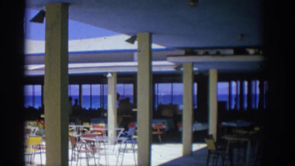 1962 Rhodes frame showing the restaurant from inside, with columns and seating silhouetted against windows overlooking the sea.