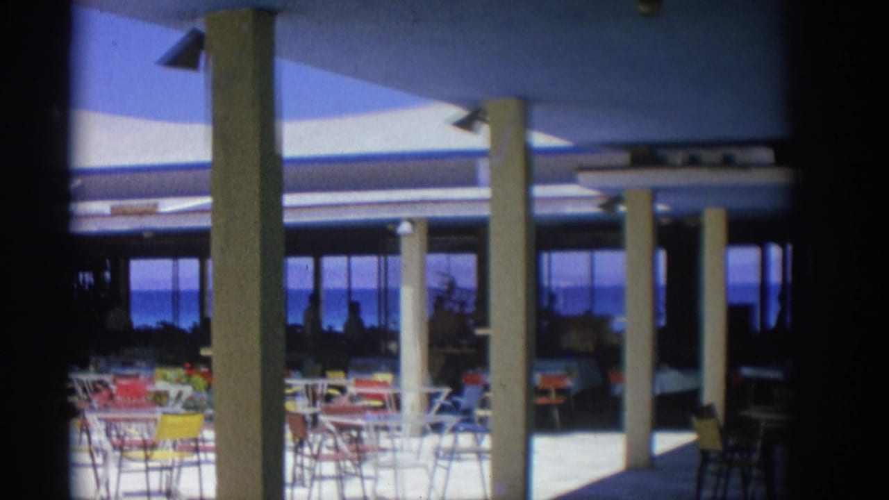 Outdoor restaurant terrace with colorful chairs, columns, and sea visible beyond glass openings.