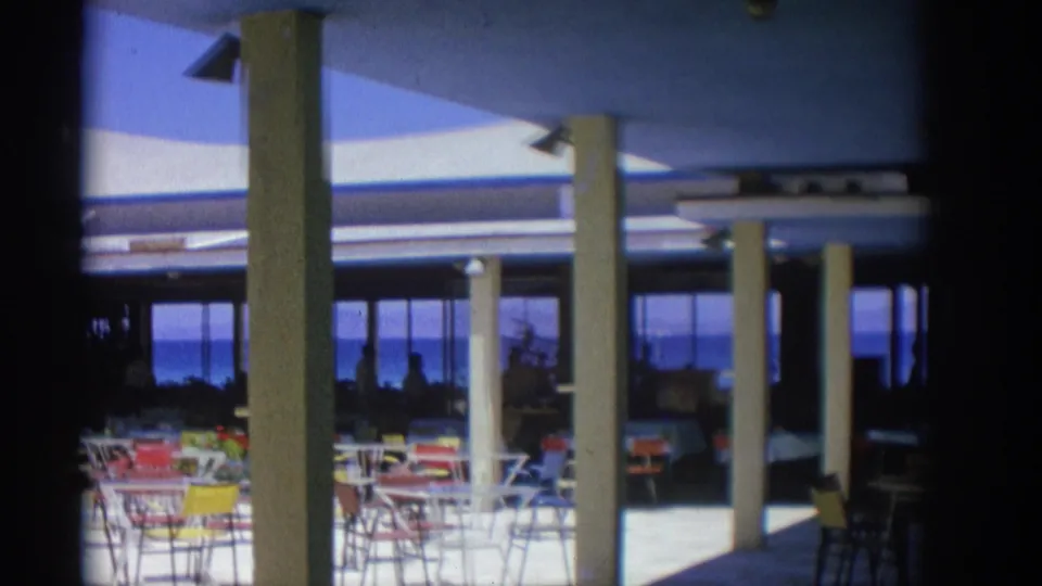 1962 Rhodes home-movie frame of a shaded seaside restaurant terrace with colorful chairs, columns, and silhouetted patrons.