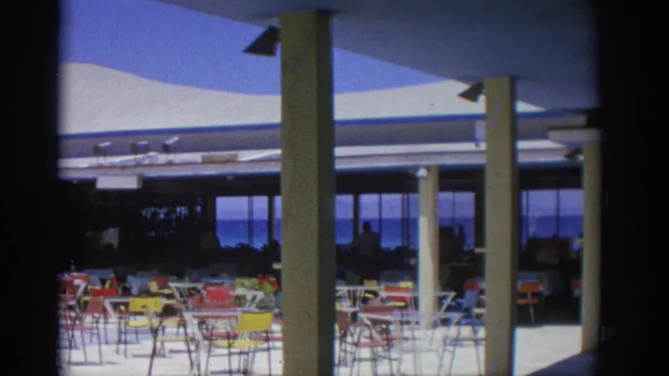 Rhodes, Greece, 1962 terrace view with colorful outdoor chairs, modern columns, and dark seated figures near the sea.