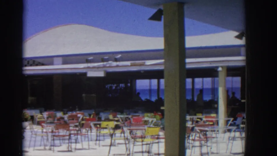 1962 Rhodes seaside restaurant terrace showing colorful chairs, planters, columns, and silhouetted people under shade.