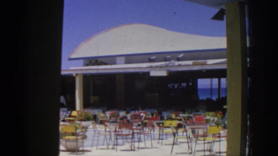 1962 Rhodes resort terrace with red and yellow patio chairs, small tables, and a modern white canopy near the coast.