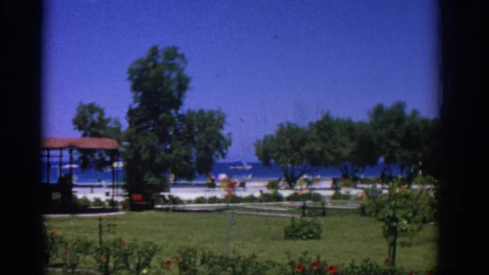 1962 Rhodes coastal resort grounds with manicured lawn, flowering beds, trees, beach activity, and sea beyond.