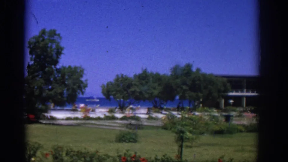 Rhodes resort garden scene showing lawn, trees, flowers, beachgoers, sea, and part of a modern building at frame edge.