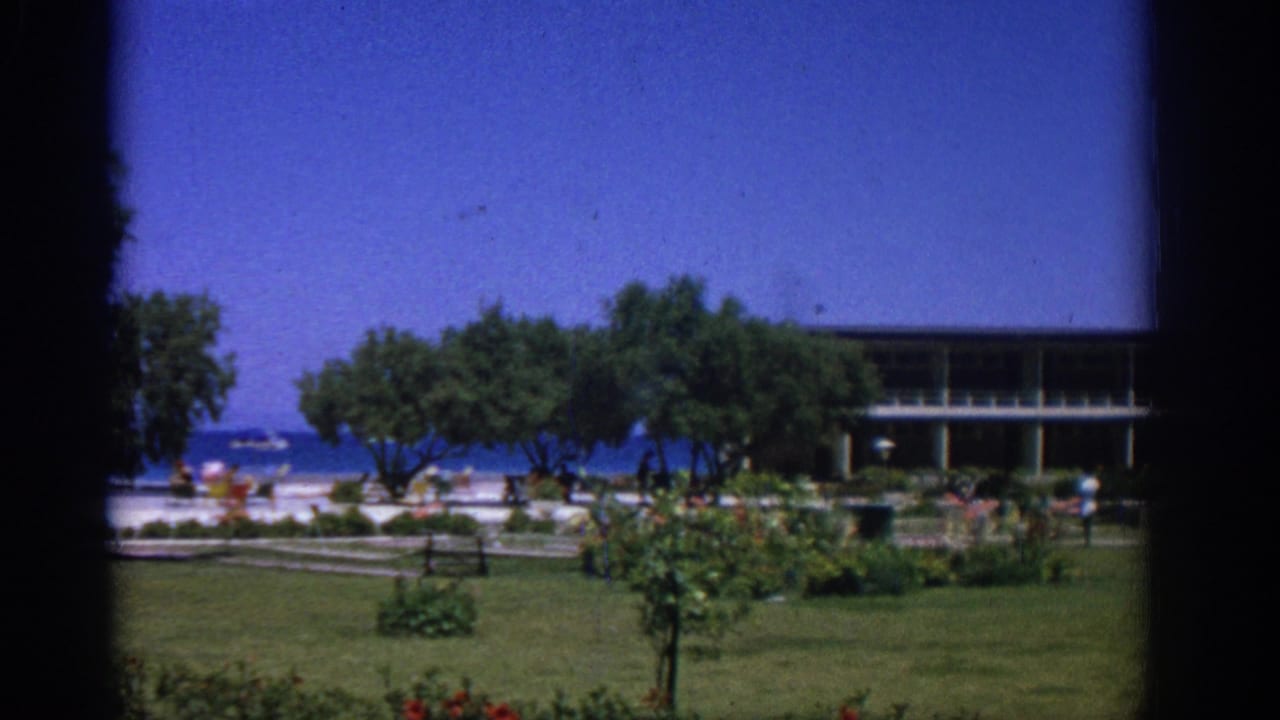 1962 Rhodes 8mm frame looking across a green garden toward a low modern building, trees, blue sea, and small distant people near the shore.