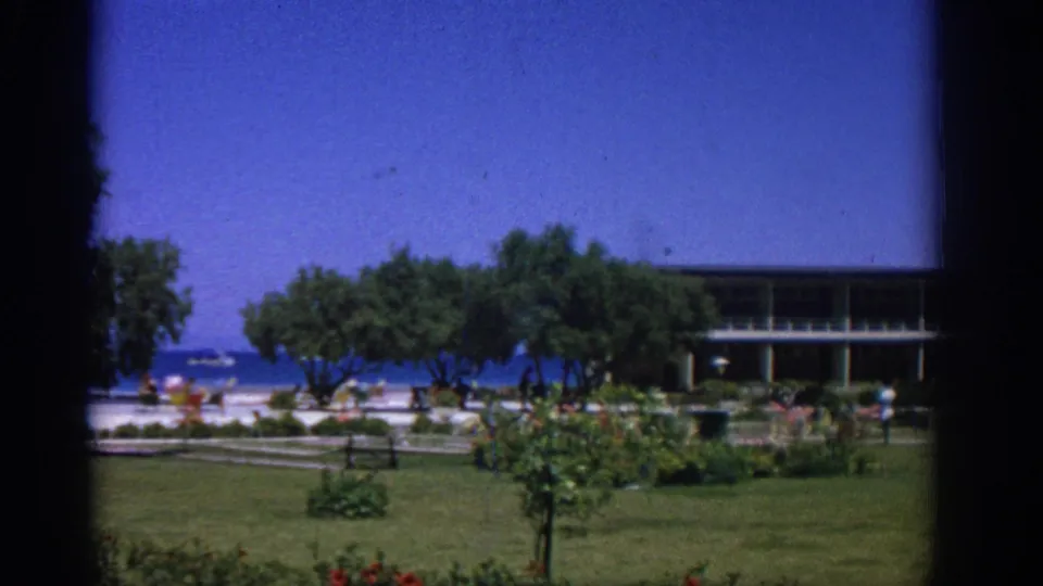 1962 Rhodes 8mm frame looking across a green garden toward a low modern building, trees, blue sea, and small distant people near the shore.