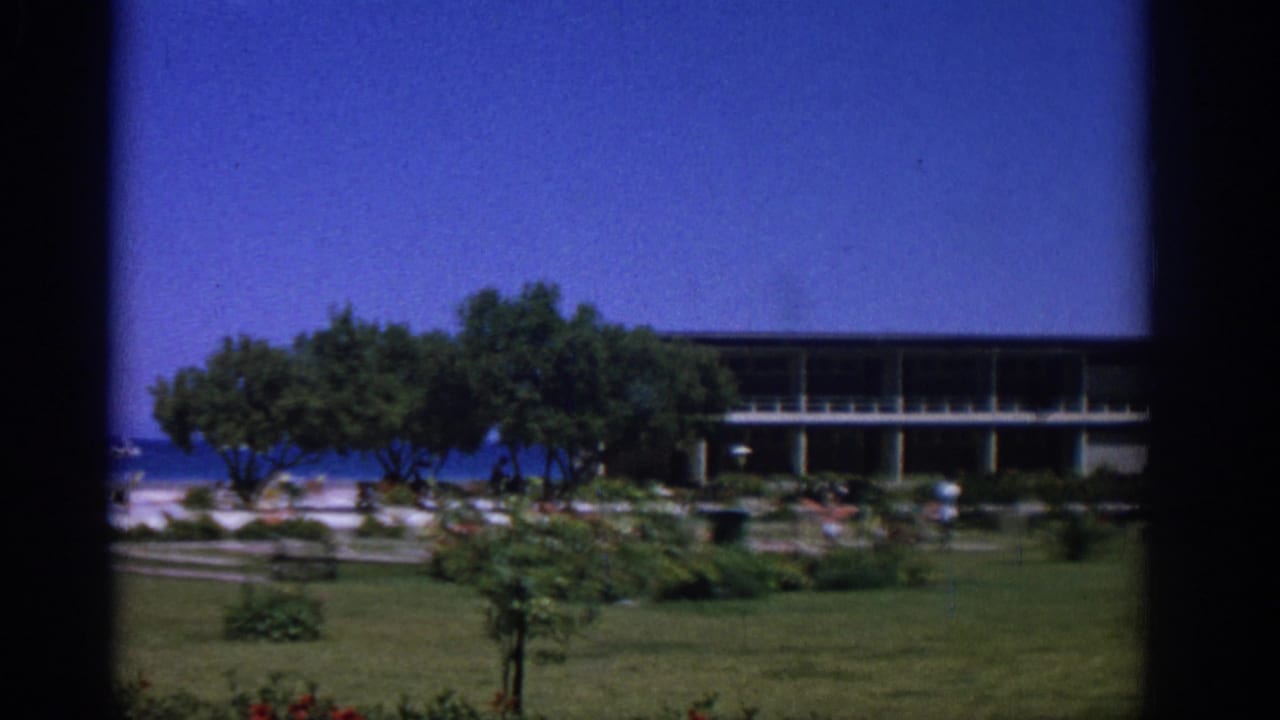 1962 Rhodes travel frame with a garden foreground, shaded trees, a long balcony building, beachgoers, blue sea, and a distant boat.