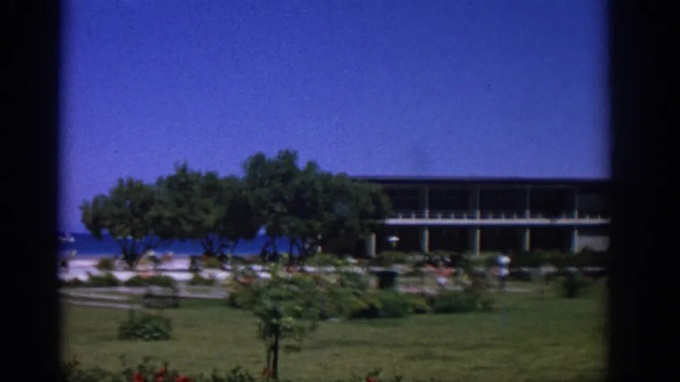 1962 Rhodes travel frame with a garden foreground, shaded trees, a long balcony building, beachgoers, blue sea, and a distant boat.