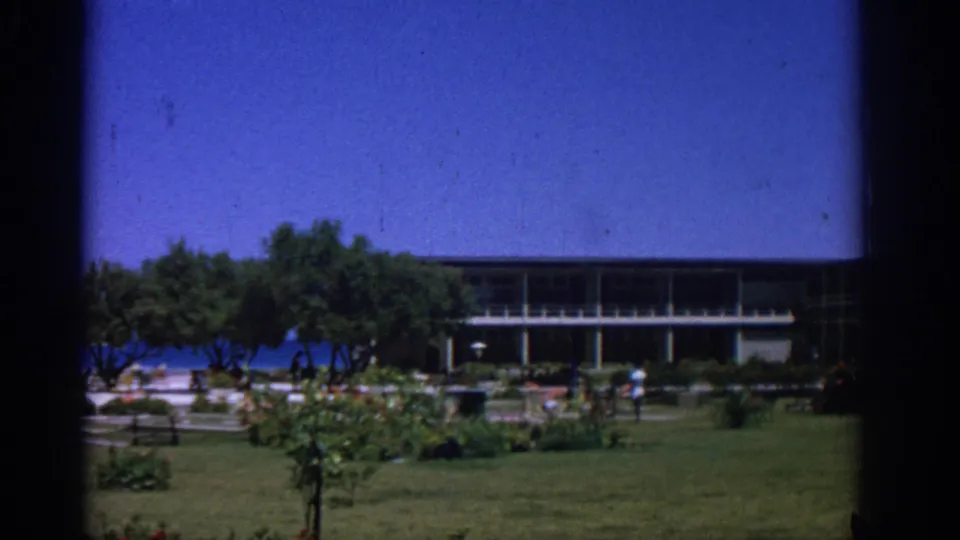 Rhodes 1962 home-movie frame showing a lawn and planted garden before a low seaside building, with blue water visible through the trees.