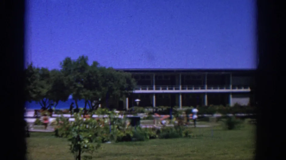 Late-frame 1962 Rhodes view across a sunny garden toward the grey balcony building, with trees, lawn, sea, and distant beach visitors.