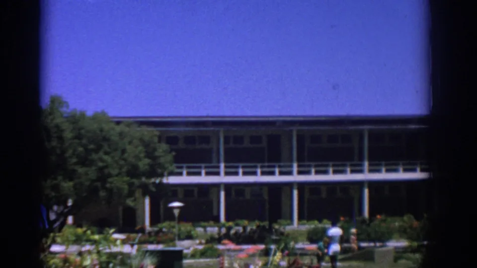 1962 Rhodes frame with a landscaped garden, balcony-fronted building, trees and small distant figures in bright daylight.