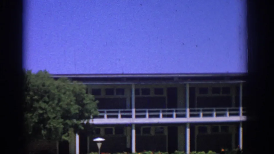 1962 Rhodes home-movie view of a mid-century building facade above a garden, framed by trees and film gate edges.