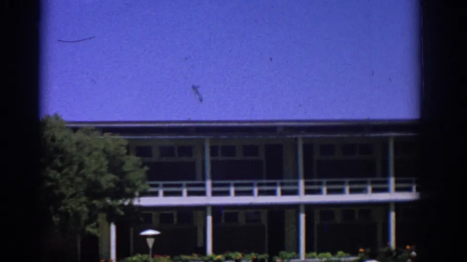 Rhodes 1962 frame showing the garden-front building, white balcony railings, a lamp post, trees and saturated blue sky.