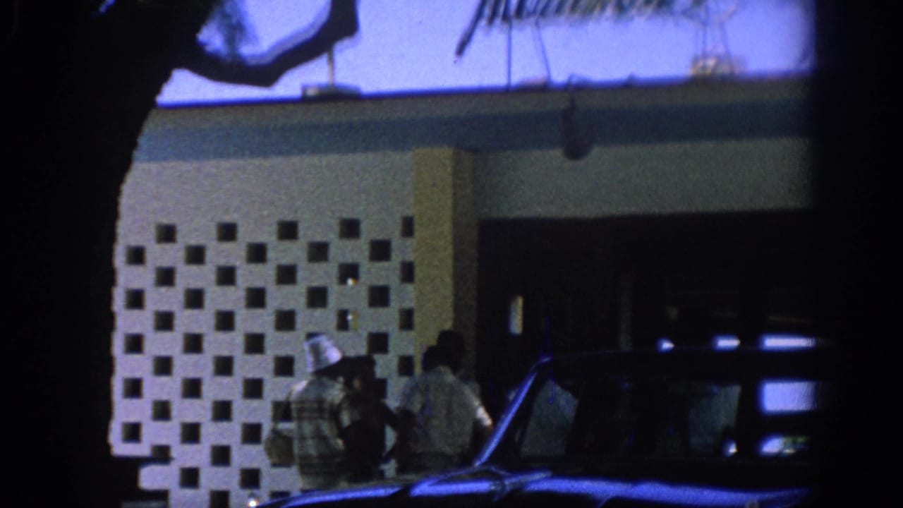 People stand near a patterned building facade with a dark car in front.