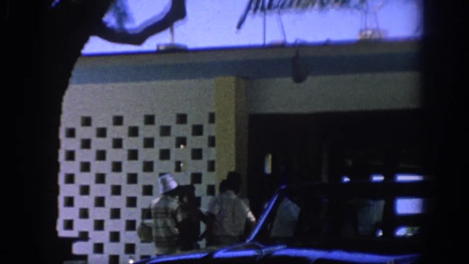 1962 Rhodes home-movie frame showing people near a geometric building entrance with a dark classic car partially in view.