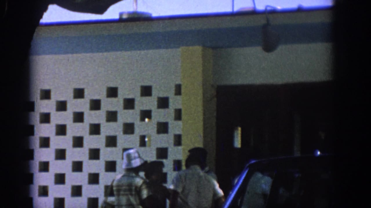 People stand by a patterned building entrance beside a dark car.